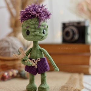 May include: A green crocheted doll with purple hair and purple shorts, holding a small beige slingshot. The doll has black button eyes, a stitched smile, and the text @VERMA.TOYS is visible on the doll's torso. The figure is crafted with yarn.