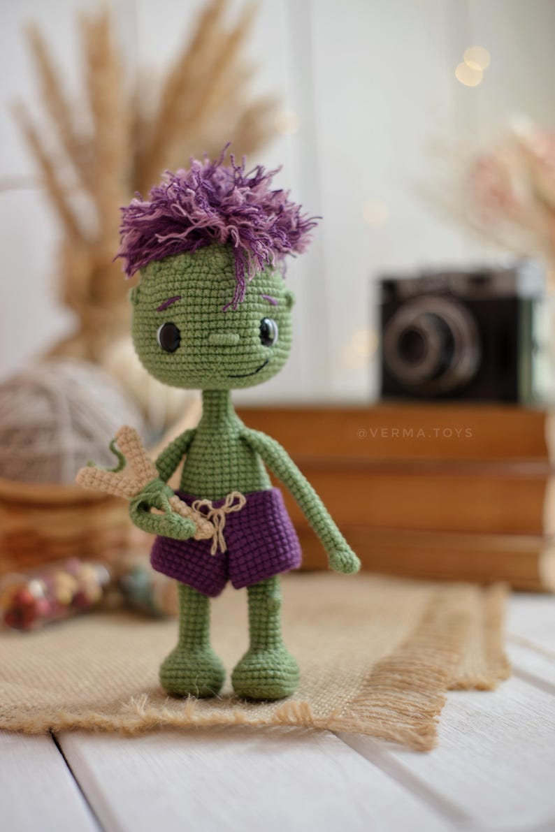 May include: A green crocheted doll with purple hair and purple shorts, holding a small beige slingshot. The doll has black button eyes, a stitched smile, and the text @VERMA.TOYS is visible on the doll's torso. The figure is crafted with yarn.