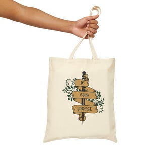 May include: Cream tote bag featuring a sword design piercing a banner with the words "Je Suis Prest" in brown. Green floral elements surround the banner. The bag is held by a person.