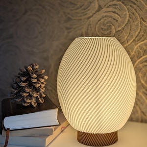 Puede incluir: Una lámpara crema en forma de huevo con un diseño en espiral, colocada sobre una base marrón. Una piña y libros apilados con un marcador están junto a la lámpara. La lámpara está sobre una superficie blanca.