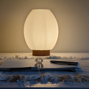 Puede incluir: Una lámpara ovalada y brillante con base de madera. La pantalla tiene un diseño en espiral. Un cuaderno y un bolígrafo están en primer plano, sugiriendo un espacio de trabajo o una mesita de noche. El ambiente es cálido.