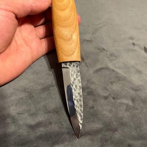 Puede incluir: Un peque&ntilde;o cuchillo artesanal con un mango de madera clara y una hoja de metal martillado. La hoja es plateada y termina en punta afilada. El mango es liso y redondeado, mostrando la veta de la madera.