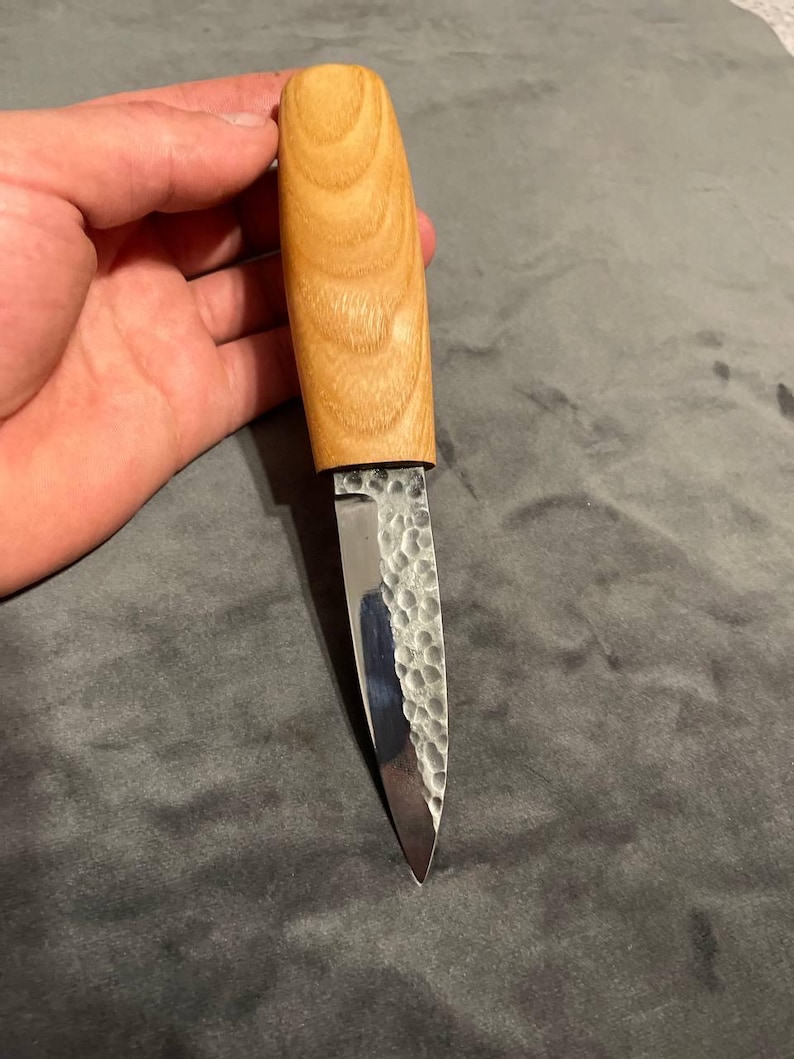 Puede incluir: Un peque&ntilde;o cuchillo artesanal con un mango de madera clara y una hoja de metal martillado. La hoja es plateada y termina en punta afilada. El mango es liso y redondeado, mostrando la veta de la madera.