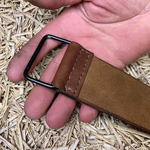 May include: A close-up of a brown leather belt with a black metal buckle. The belt is held by a hand, showcasing the texture and stitching details. The background is a textured surface.