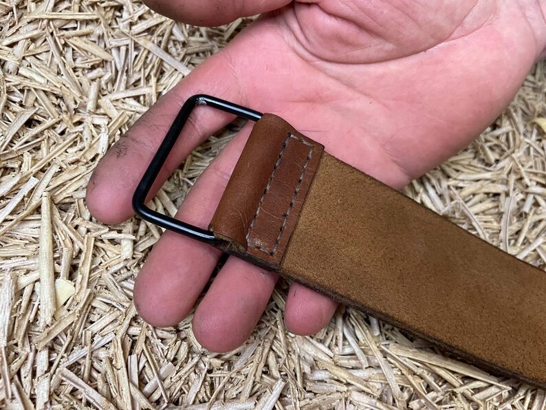 May include: A close-up of a brown leather belt with a black metal buckle. The belt is held by a hand, showcasing the texture and stitching details. The background is a textured surface.