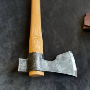 Könnte beinhalten: Eine handgefertigte Axt mit einem hellen Holzgriff und einem dunklen Metallkopf. Der Axtkopf hat eine scharfe, abgeschrägte Kante. Der Griff ist glatt und scheint aus Hartholz zu sein. Eine Lederscheide ist im Hintergrund sichtbar.