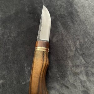 Può includere: Un coltello artigianale con una lama curva in acciaio inossidabile. L'impugnatura è in legno scuro con un motivo a venature naturali, impreziosita da un anello in ottone. Il coltello è progettato per tagliare e affettare.