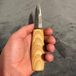 Puede incluir: Un peque&ntilde;o cuchillo artesanal con un mango de madera clara y una hoja plateada. La hoja tiene un aspecto texturizado y martillado. El cuchillo se sostiene en una mano sobre un fondo gris.