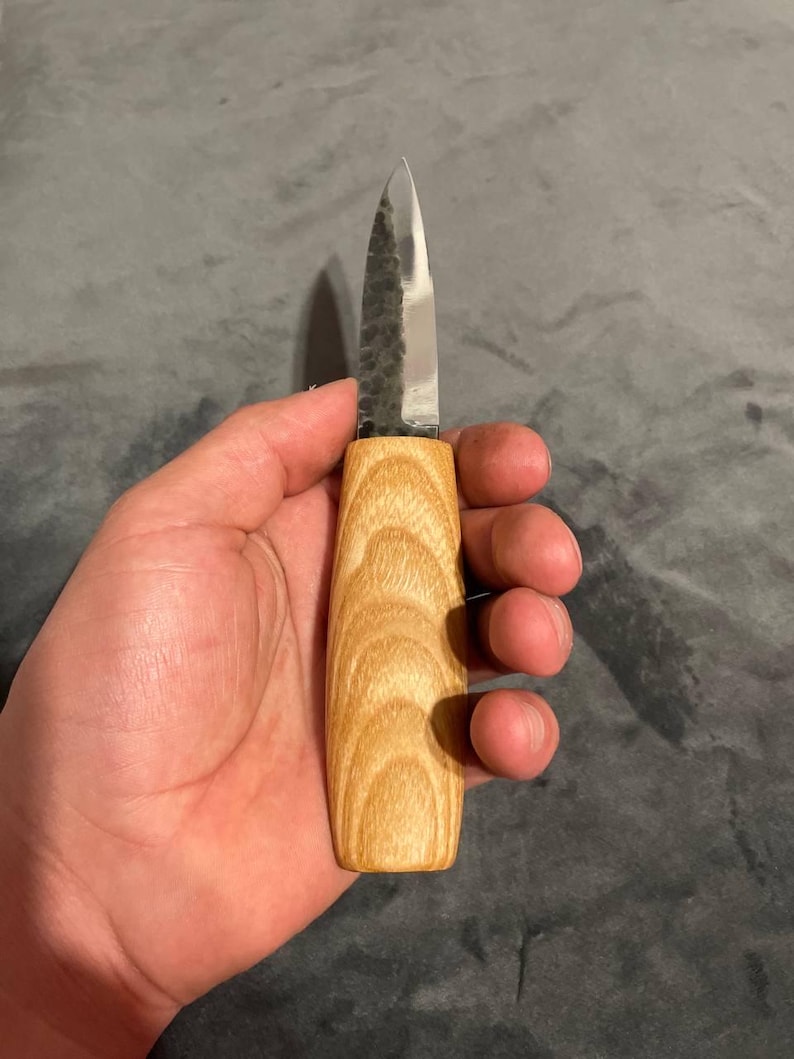 Puede incluir: Un peque&ntilde;o cuchillo artesanal con un mango de madera clara y una hoja plateada. La hoja tiene un aspecto texturizado y martillado. El cuchillo se sostiene en una mano sobre un fondo gris.