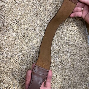 May include: A brown leather strop with a black metal loop at one end and a stitched handle at the other. The strop is curved and appears to be used for sharpening straight razors. The background is a bed of straw.