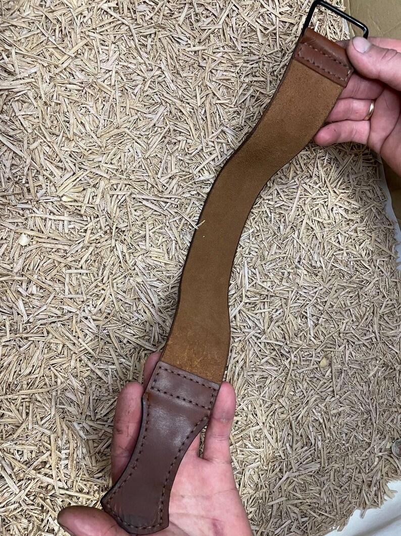 May include: A brown leather strop with a black metal loop at one end and a stitched handle at the other. The strop is curved and appears to be used for sharpening straight razors. The background is a bed of straw.