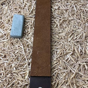 Puede incluir: Un cuero de afeitar de cuero marrón con un mango marrón oscuro y una arandela plateada. Una pequeña barra rectangular de compuesto de afilado azul claro está al lado del cuero. Los artículos están sobre un fondo de virutas de madera.