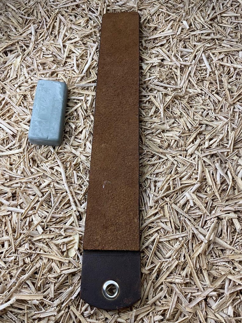 May include: A brown leather strop with a dark brown handle and a silver grommet. A small, rectangular, light blue sharpening compound bar is next to the strop. The items are on a background of wood shavings.