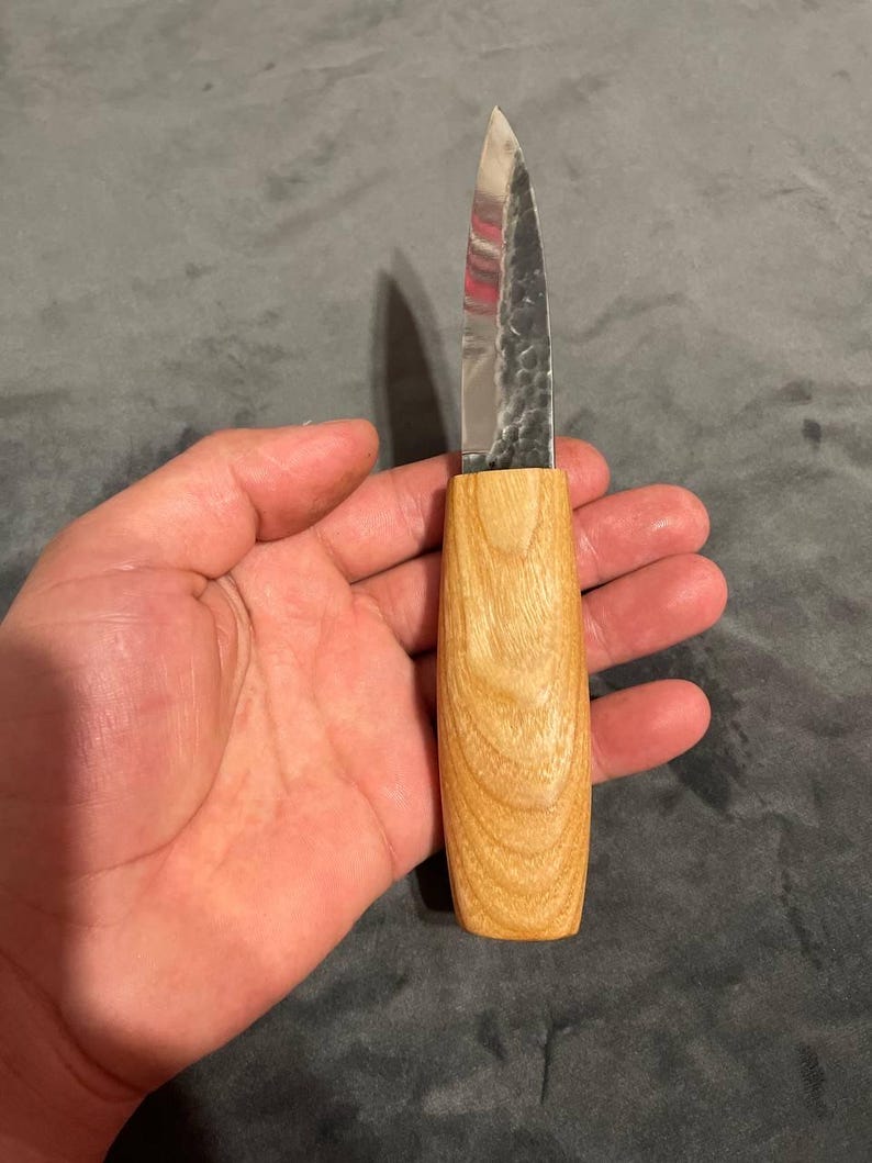 Puede incluir: Un peque&ntilde;o cuchillo artesanal con un mango de madera clara y una hoja plateada. La hoja tiene una superficie texturizada. El cuchillo est&aacute; dise&ntilde;ado para tareas de corte de precisi&oacute;n.