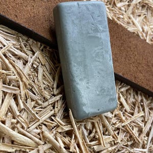 Puede incluir: Una piedra de afilar rectangular, de color gris claro, descansa sobre una correa de cuero marrón, sobre un fondo de virutas de madera. La superficie de la piedra es ligeramente texturizada. La correa de cuero es de un marrón oscuro intenso.