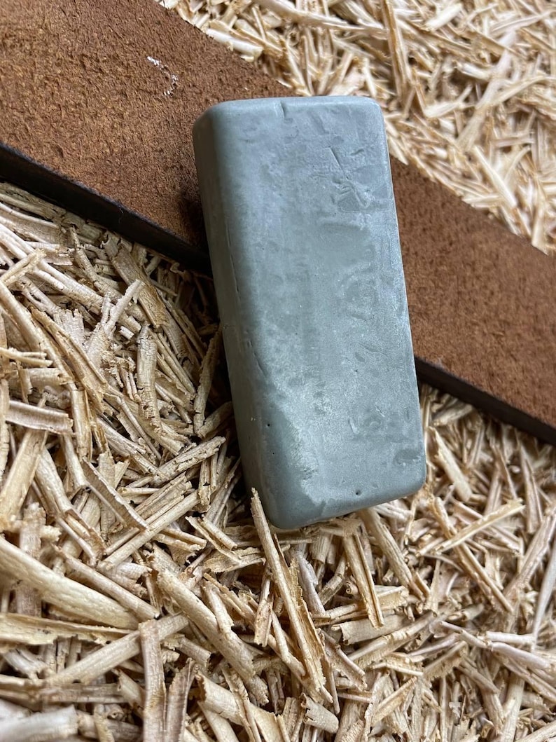 May include: A rectangular, light gray sharpening stone rests on a brown leather strap, set against a background of wood shavings. The stone's surface appears slightly textured. The leather strap is a rich, dark brown.