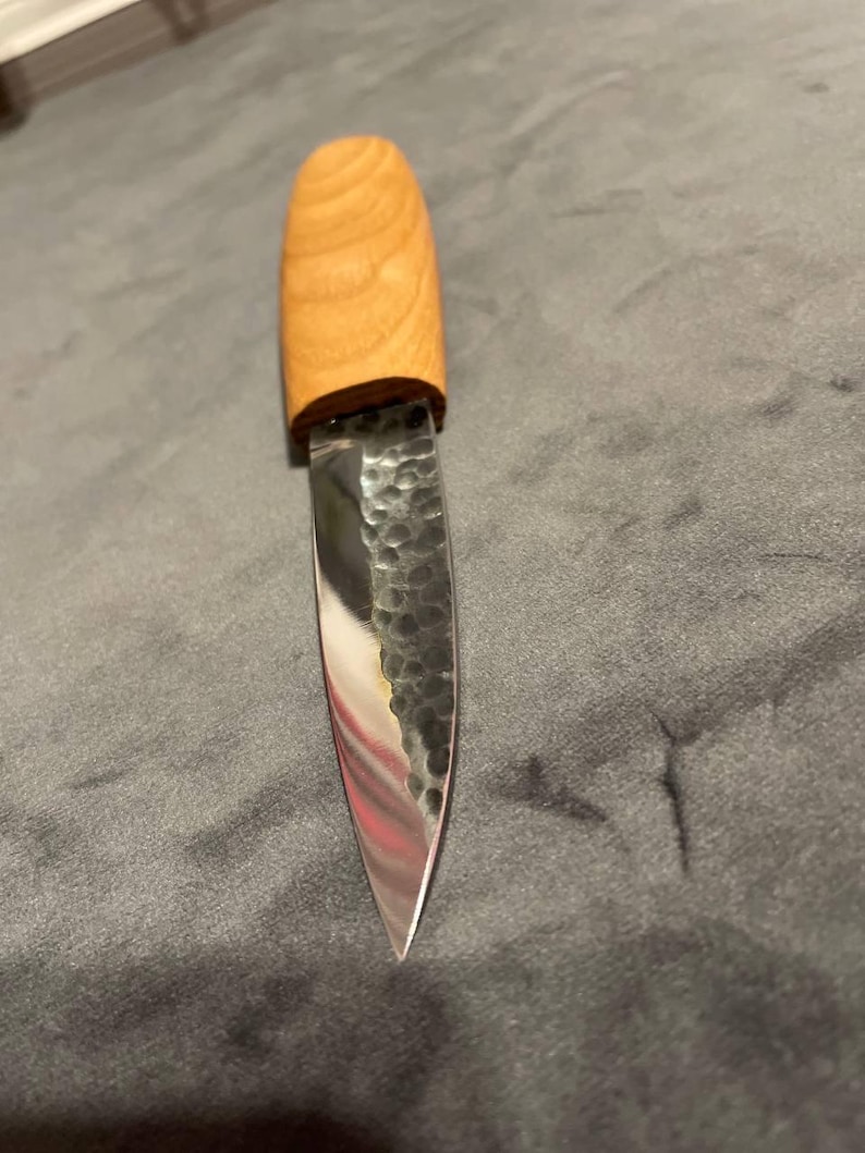 Puede incluir: Un peque&ntilde;o cuchillo artesanal con un mango de madera clara y una hoja de metal martillado. La hoja tiene una punta afilada y una superficie reflectante. El cuchillo est&aacute; dise&ntilde;ado para tareas de corte de precisi&oacute;n.