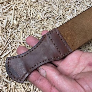 May include: A close-up of a brown leather strop with a stitched handle. The strop is made of a lighter brown leather and is held in a hand. The background is a light brown, textured surface.