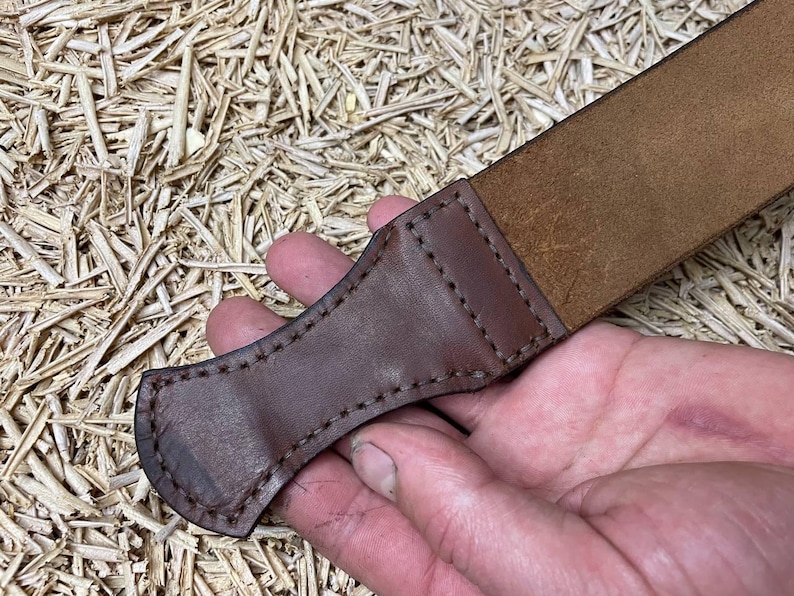 May include: A close-up of a brown leather strop with a stitched handle. The strop is made of a lighter brown leather and is held in a hand. The background is a light brown, textured surface.