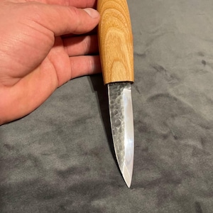 Puede incluir: Un cuchillo artesanal con un mango de madera clara y una hoja plateada. La hoja tiene una superficie texturizada. El cuchillo se sostiene en una mano.