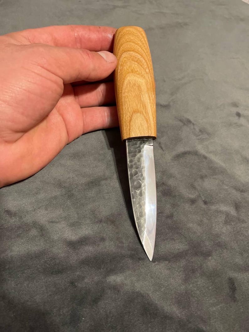 Puede incluir: Un cuchillo artesanal con un mango de madera clara y una hoja plateada. La hoja tiene una superficie texturizada. El cuchillo se sostiene en una mano.