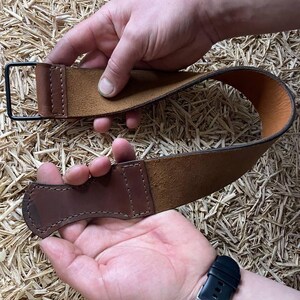 May include: A brown leather strop with a dark brown handle and a black metal loop. The strop is held up by two hands, showcasing the leather's texture and craftsmanship. The background is a textured surface.