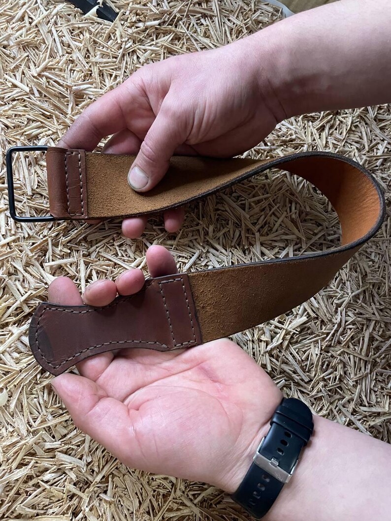 May include: A brown leather strop with a dark brown handle and a black metal loop. The strop is held up by two hands, showcasing the leather's texture and craftsmanship. The background is a textured surface.