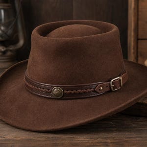 May include: A dark brown felt hat with a flat crown and a wide brim. The hat features a dark brown leather band decorated with a braided leather strip, a circular metallic concho ornament, and a small adjustable metal buckle on the side.