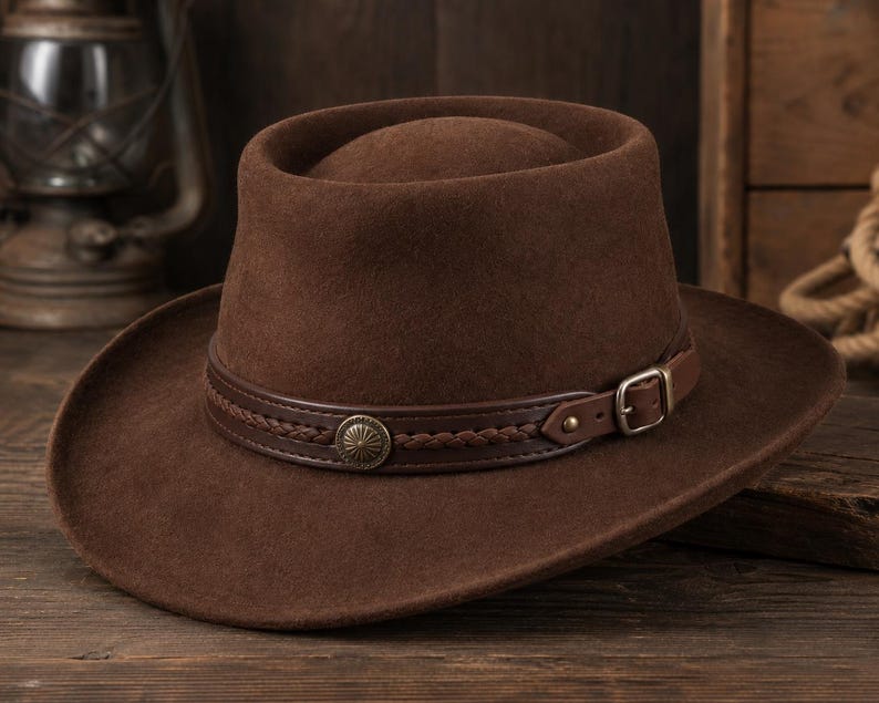 May include: A dark brown felt hat with a flat crown and a wide brim. The hat features a dark brown leather band decorated with a braided leather strip, a circular metallic concho ornament, and a small adjustable metal buckle on the side.