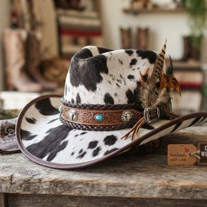 May include: A cowprint western hat featuring a brown leather band with braided edges, silver-toned metal accents, and turquoise stones. The hat is adorned with a cluster of feathers on the side and sits next to a tag labeled COWPRINT WESTERN TOP HAT.