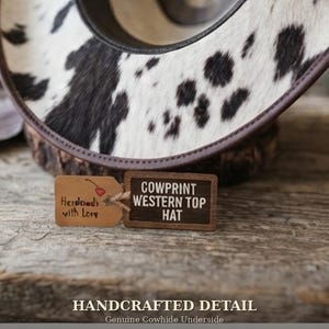 May include: A western top hat featuring a black and white cow print pattern with a dark brown brim edge. Attached are two small rectangular tags, one brown with a cherry illustration and text, and one dark wood with the text COWPRINT WESTERN TOP HAT.