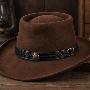 Può includere: Un cappello in feltro marrone con corona piatta e tesa larga. Il cappello presenta una fascia in pelle nera decorata con un motivo intrecciato, un concho metallico circolare centrale e una piccola fibbia color ottone sul lato.