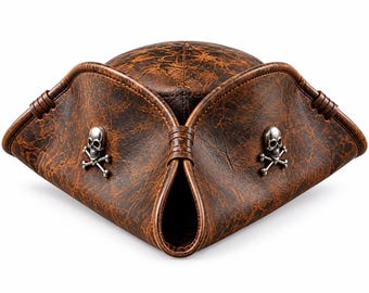 May include: A brown tricorn hat with a distressed, weathered texture. The hat features three upturned brims secured with stitched detailing and two silver-toned metal skull and crossbones emblems attached to the front-facing panels.