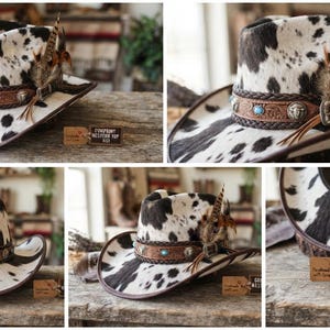 May include: A western hat featuring a black and white cow print pattern. The hat is adorned with a brown leather band, a turquoise stone, silver-toned metal accents, and a cluster of brown and tan feathers. A small tag reads Cowprint Western Top Hat.