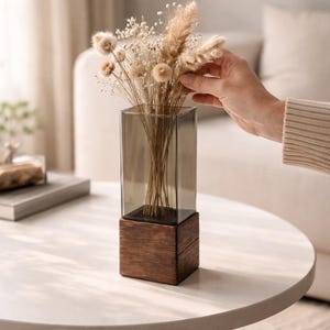May include: A modern decorative vase with a rectangular, smoky glass top and a dark wooden base. The vase holds a bouquet of dried flowers and grasses. The vase is sitting on a white table.