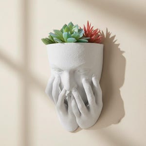 Peut inclure: Pot mural blanc en forme de visage avec les mains couvrant les yeux et la bouche. Le pot contient des succulentes artificielles vertes et rouges. La surface texturée du pot contraste avec les plantes lisses.