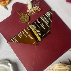 May include: A deep burgundy envelope containing a gold-toned acrylic invitation card with white text. The card is secured with a gold wax seal, gold metallic thread, and a small arrangement of dried white flowers and grasses.