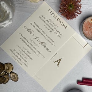 May include: Ivory wedding invitation with gold lettering and a monogram. The invitation is surrounded by floral accents, including a cream-colored rose and a small, spiky flower. Gold wax seals and candles are also present.