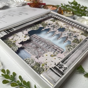 May include: A white, layered invitation box featuring a detailed architectural illustration with floral accents. The box has a decorative border and a lid, creating a 3D effect. The design incorporates shades of white, gray, and pastel colors.