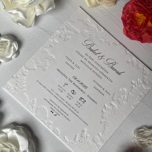 Puede incluir: Una invitación de boda blanca en relieve con diseños florales y de hojas. La invitación incluye los nombres "Buket & Burak" y los detalles del evento. La invitación está rodeada de rosas blancas, crema y rojas.