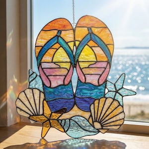 May include: Stained glass window art featuring a pair of flip-flops, seashells, and starfish. The artwork uses vibrant colors like orange, yellow, blue, and pink to depict a beach scene. The piece is designed to hang and is displayed in front of a window.