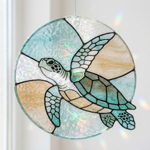 May include: A stained glass suncatcher featuring a sea turtle design. The turtle is depicted in shades of teal and white, set against a backdrop of light blue and tan glass. The suncatcher is round and hangs from a silver chain.