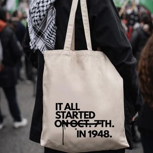 Könnte beinhalten: Beige Tragetasche mit schwarzem Text: "IT ALL STARTED ON OCT. 7TH. IN 1948." Die Tasche wird von einer Person getragen, die eine schwarze Jacke und einen schwarz-weiß karierten Schal trägt. Im Hintergrund sind Flaggen zu sehen.