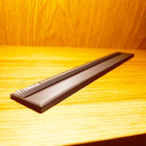 May include: A dark grey, rectangular object with a recessed channel. The text "17-20mm" is printed in white. It is placed on a wooden surface. The channel appears to be for holding a small item.