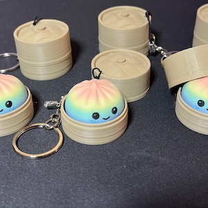 May include: A collection of keychains featuring a small, rainbow-coloured dumpling with a smiling face, housed inside a tan, cylindrical, two-piece steamer basket container. Each keychain includes a metal ring for attachment.