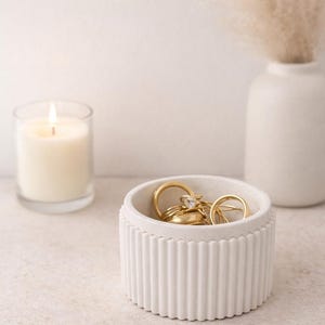 Könnte beinhalten: Eine weiße, geriffelte Schmuckschale gefüllt mit goldenen Ringen. Eine brennende Kerze in einem Glas und eine weiße Vase mit getrockneten Blumen befinden sich im Hintergrund. Die Szene ist auf einer neutral getönten Oberfläche aufgebaut.
