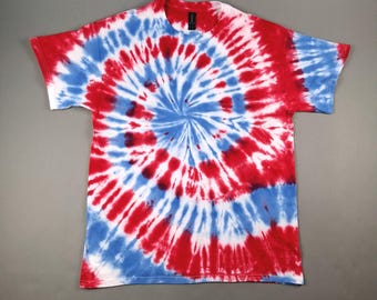May include: A short-sleeved t-shirt with a spiral tie-dye pattern in red, white, and blue. The design radiates from the center, creating a bold and colorful effect. The shirt is made of a soft material and is suitable for casual wear.