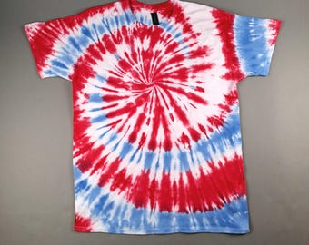 Könnte beinhalten: Ein kurzärmeliges T-Shirt mit einem spiralförmigen Batikmuster in Rot, Weiß und Blau. Das Design strahlt von der Mitte aus und erzeugt einen kräftigen und farbenfrohen Effekt. Das Shirt liegt flach auf einer grauen Oberfläche.