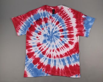 Könnte beinhalten: Ein kurzärmeliges T-Shirt mit einem spiralförmigen Batikmuster in Rot, Weiß und Blau. Das Design strahlt von der Mitte des T-Shirts aus und erzeugt einen kreisförmigen Effekt. Das Shirt ist aus einem weichen Material.