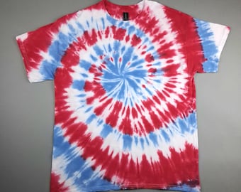 May include: A red, white, and blue tie-dye t-shirt. The design features a spiral pattern with red and blue radiating from a central white circle. The shirt has short sleeves and a crew neck.